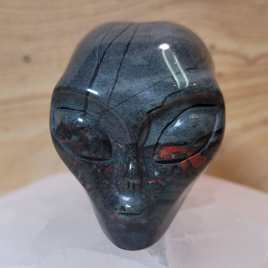 A captivating African bloodstone alien figurine, featuring rich green hues with vibrant red spots, displayed against a soft, neutral background. This unique piece symbolizes strength, courage, and curiosity, making it an intriguing decorative item.