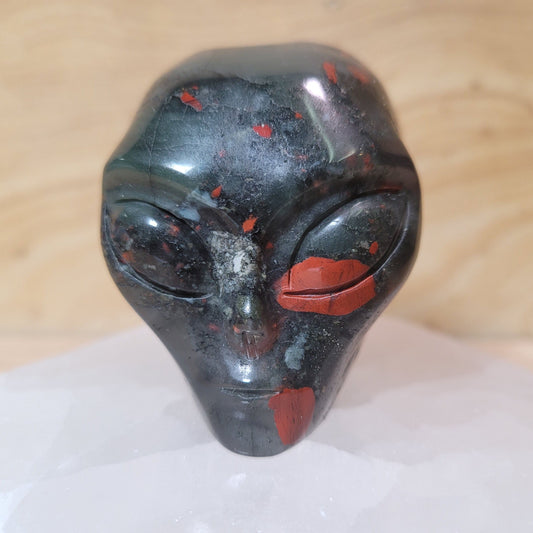 A captivating African bloodstone alien figurine, featuring rich green hues with vibrant red spots, displayed against a soft, neutral background. This unique piece symbolizes strength, courage, and curiosity, making it an intriguing decorative item.