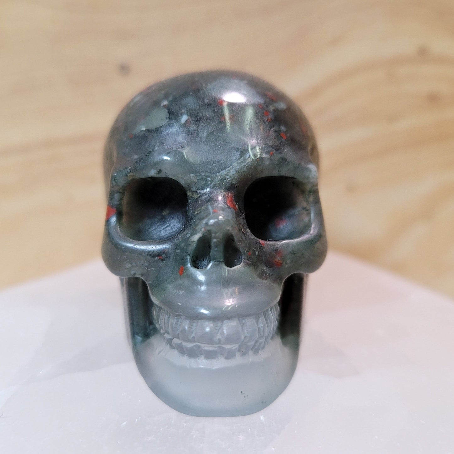 An intricately carved African bloodstone skull, showcasing rich green colors with vibrant red flecks, displayed against a soft, neutral background. This unique piece symbolizes courage, strength, and wisdom, making it an eye-catching decorative item.