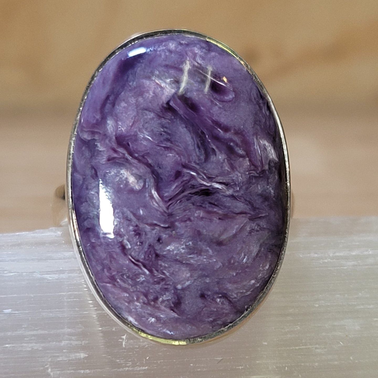 An adjustable ring featuring a polished charoite stone with vibrant purple hues and swirling patterns. The stone is set in a silver or metal band that can be adjusted to fit various finger sizes. The ring is displayed on a neutral background to showcase the stone's unique coloration and the intricate details of its pattern.