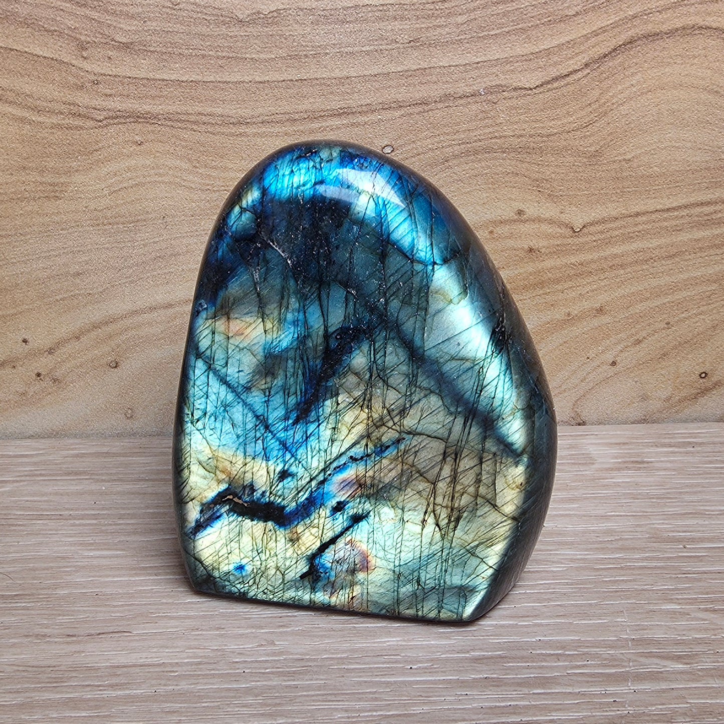 A polished labradorite stone with a natural organic shape, displaying a shimmering blue, green, and gold iridescent finish with dark inclusions, set against a light wood grain background.