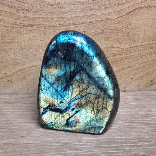 A polished labradorite stone with a natural organic shape, displaying a shimmering blue, green, and gold iridescent finish with dark inclusions, set against a light wood grain background.