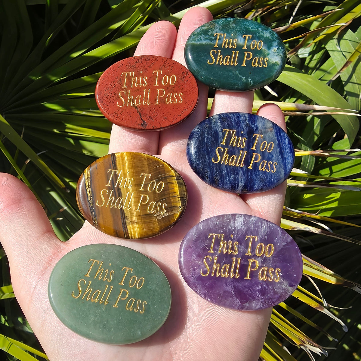Six oval polished gemstone stress stones engraved with 'This Too Shall Pass' in gold lettering, held in a person's hand against a background of green palm leaves.